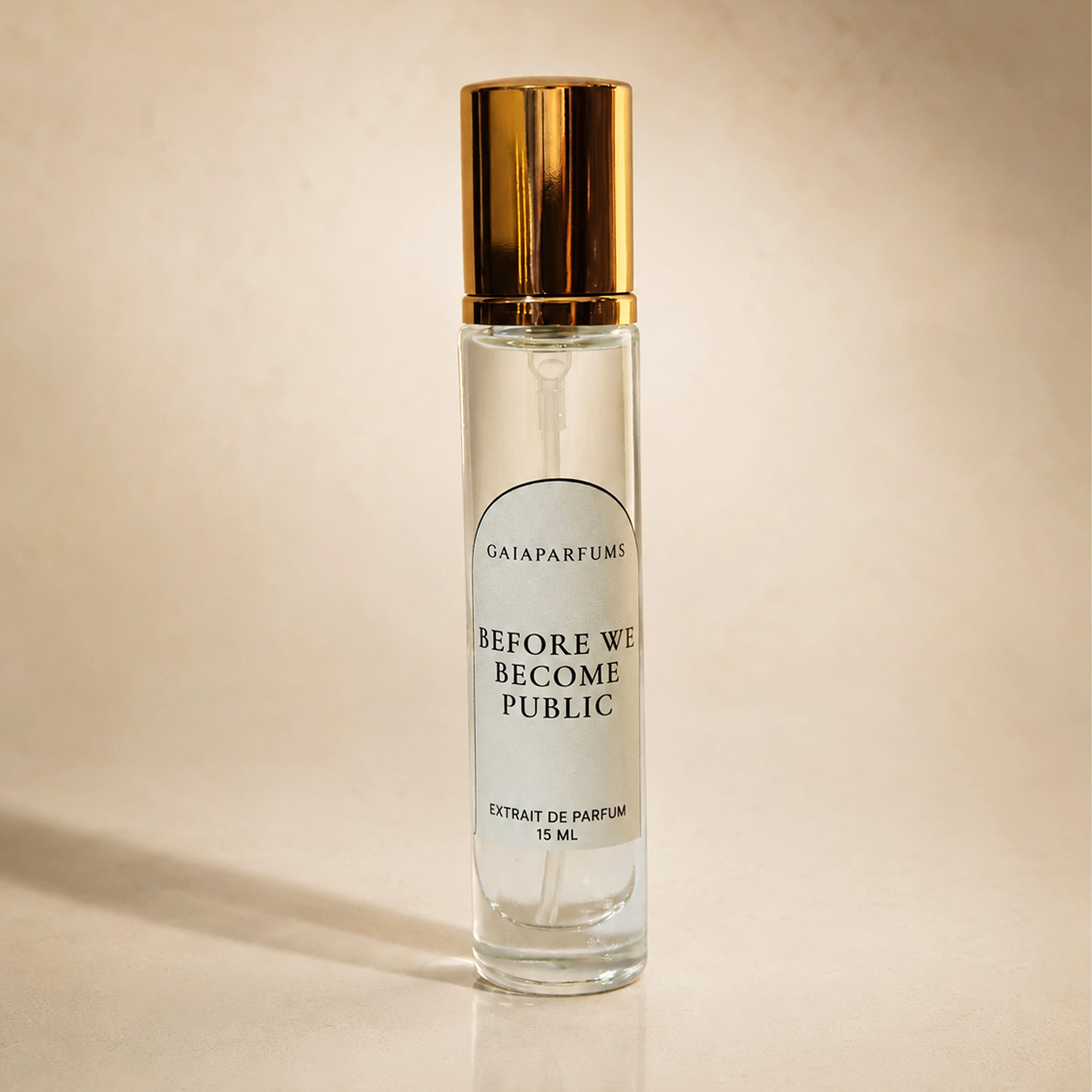 A clear glass perfume bottle with a reflective gold cap, featuring a white label that reads 'GAIAPARFUMS BEFORE WE BECOME PUBLIC EXTRAIT DE PARFUM 15 ML'. The bottle is set against a warm beige background with a soft shadow.