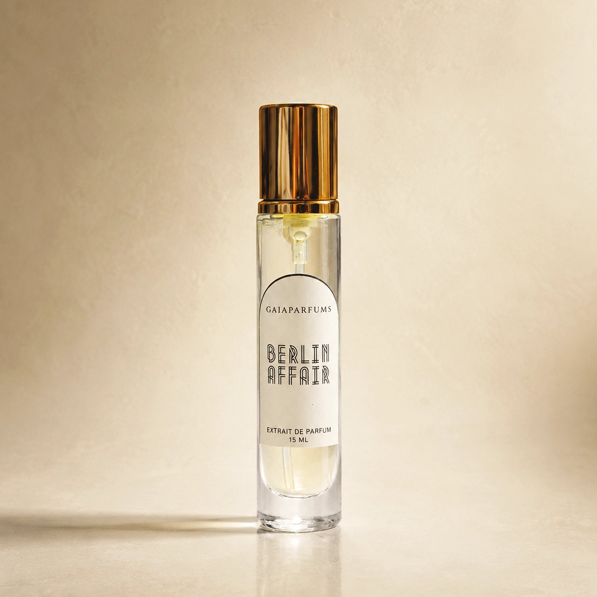 A clear glass perfume bottle with a reflective gold cap and a white label reading 'GAIA PARFUMS BERLIN AFFAIR EXTRAIT DE PARFUM 15 ML', set against a warm beige textured background.