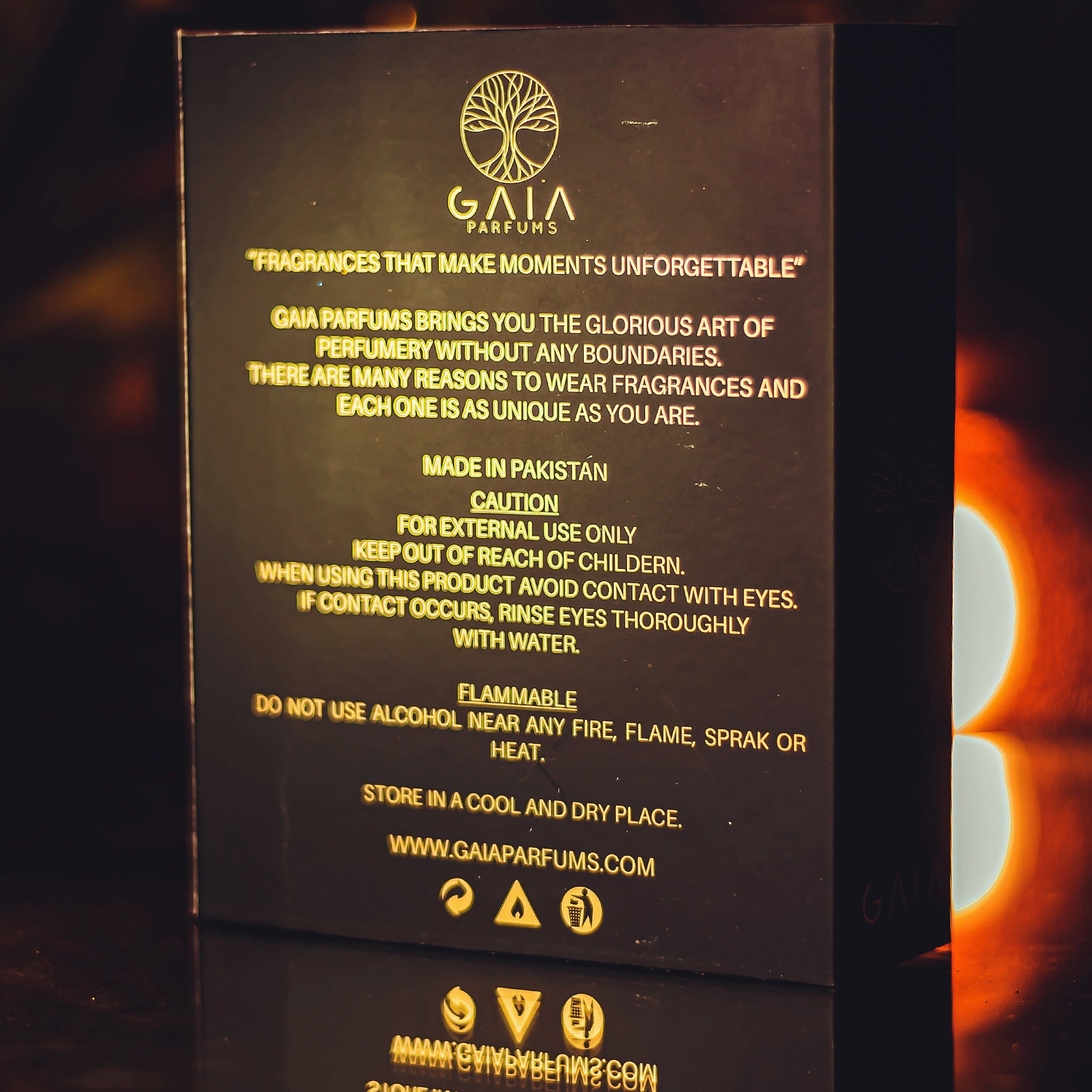 A black rectangular product information card with gold text for Gaia Parfums, showing warning and care instructions, photographed against a dark background with a warm glowing light on the right.