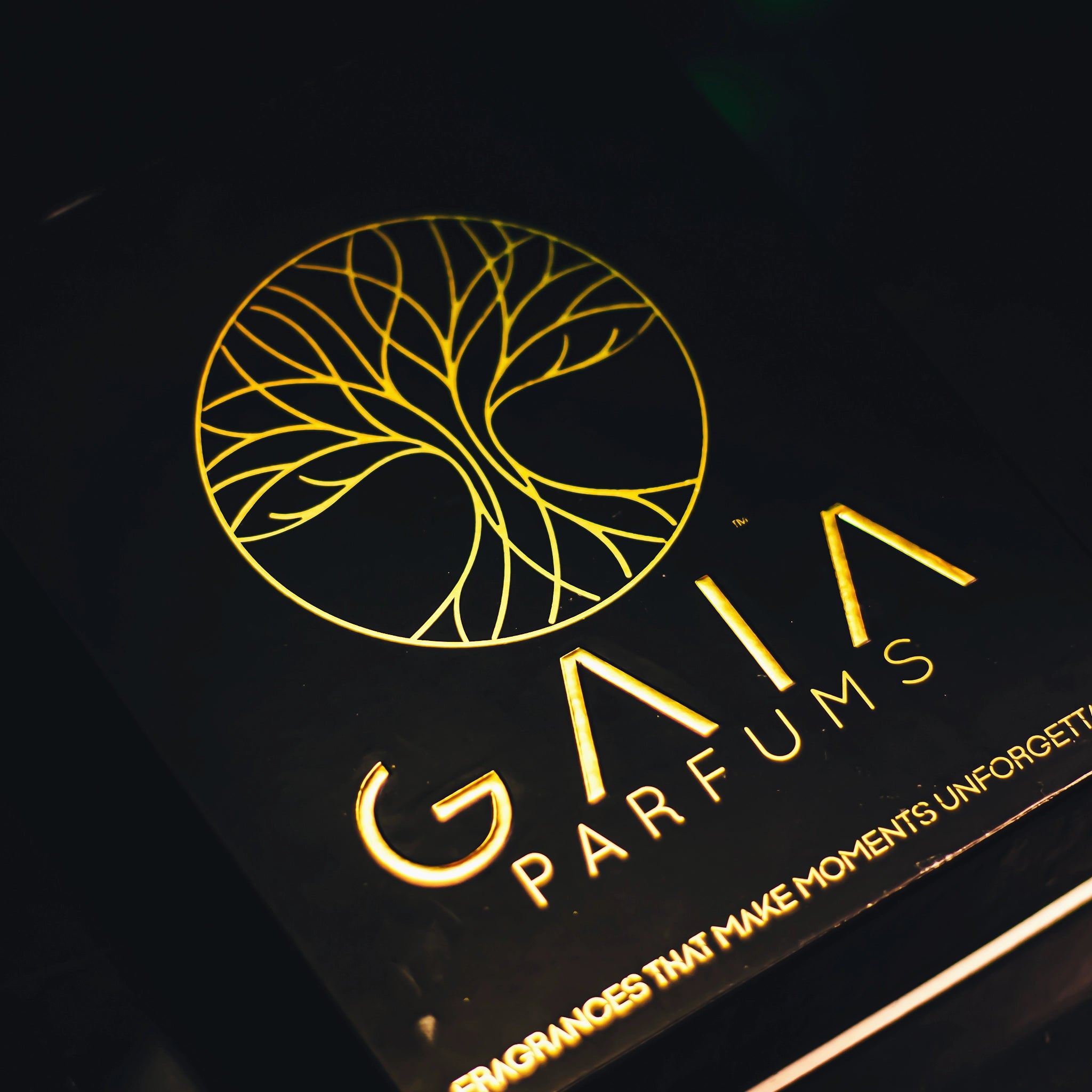 Close-up of a black box or package with gold foil branding reading 'GAIA PARFUMS' and a gold tree-of-life emblem on a dark background.