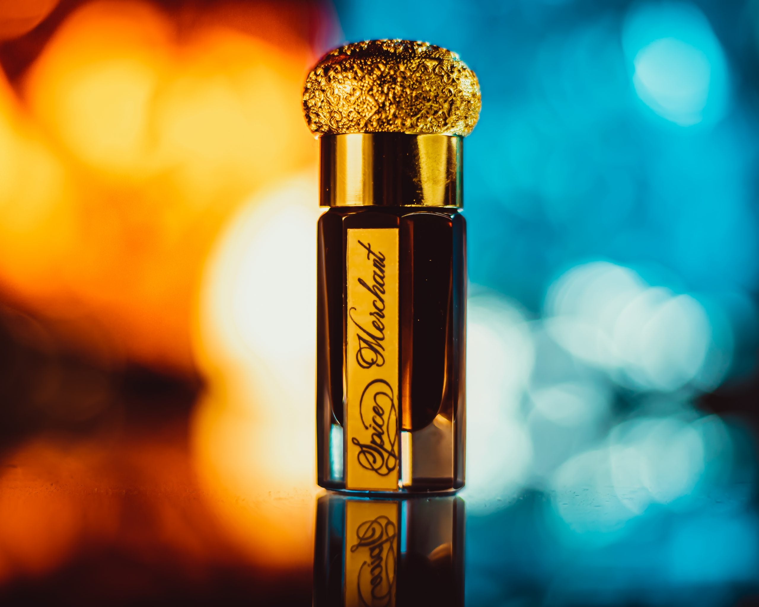 Perfume bottle with gold cap on a reflective surface against a blurred multicolored background. Spice Merchant Attar by GAIA Parfums, a Silk Route-inspired natural attar with Indian spices, saffron, rose, resins, honey and leather